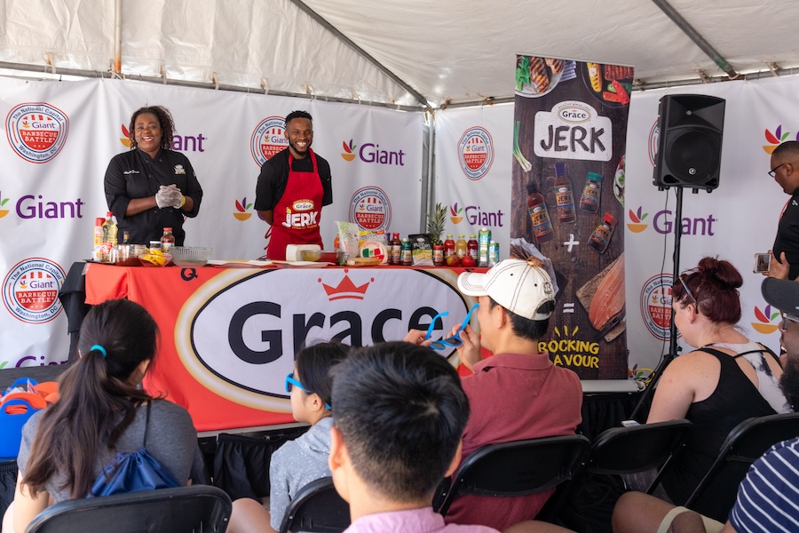 Cooking Demonstration: Two chefs, one male and one female, conducting a cooking demonstration at a BBQ event, with a crowd of spectators watching from shaded seating.