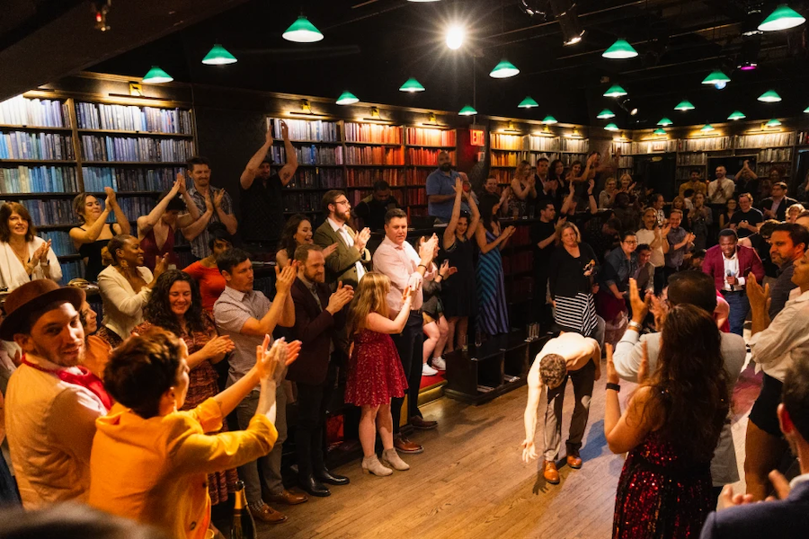  A lively crowd in a warmly lit venue applauds a performer who is taking a bow in the center of the room, surrounded by shelves of colorful books.