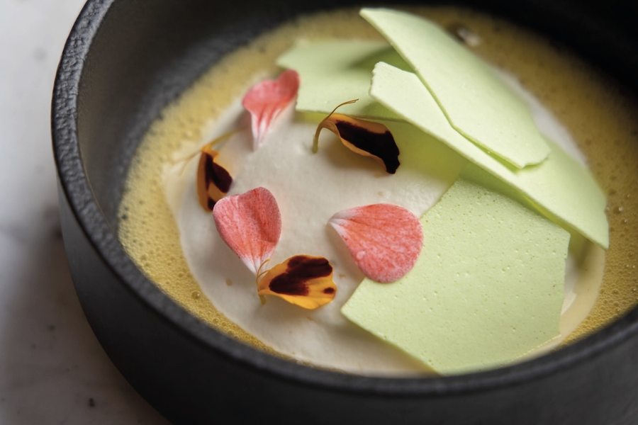 A gourmet dish in a black bowl, featuring light green shards, pink and yellow flower petals, and a creamy base.