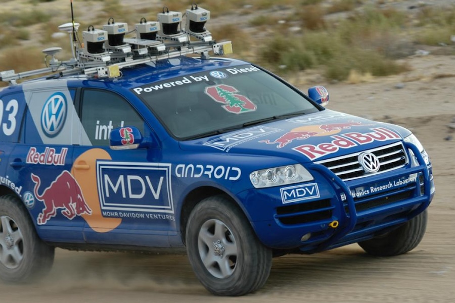 A blue Volkswagen car equipped with multiple cameras and sensors on its roof, displaying logos of various sponsors including Red Bull, MDV, and Android, driving on a dirt road.
