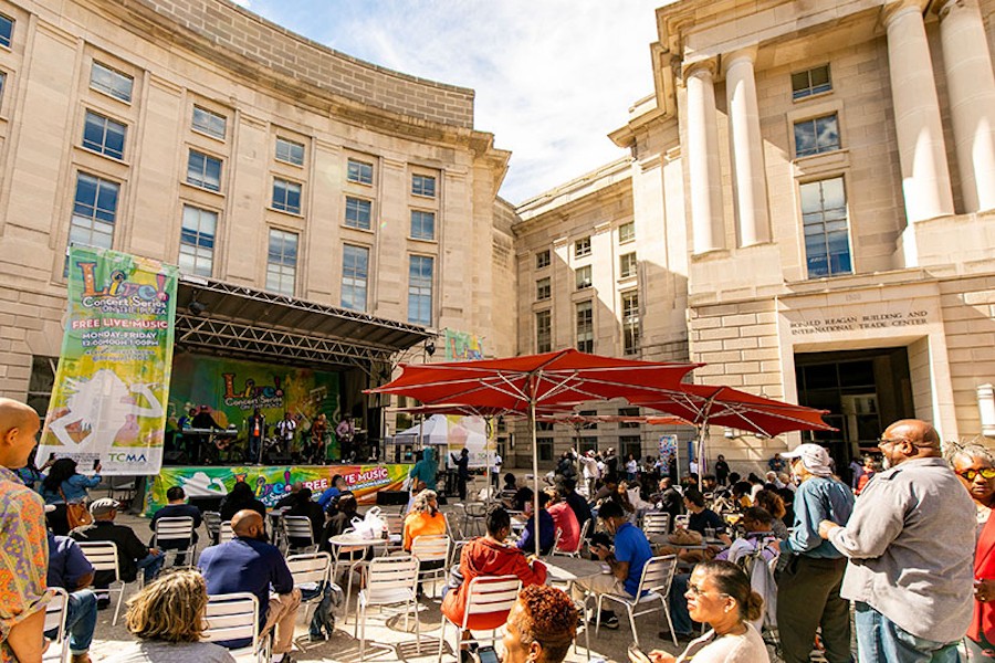 An outdoor concert taking place at the Ronald Reagan Building with a crowd of people seated and standing, enjoying live music under a sunny sky.