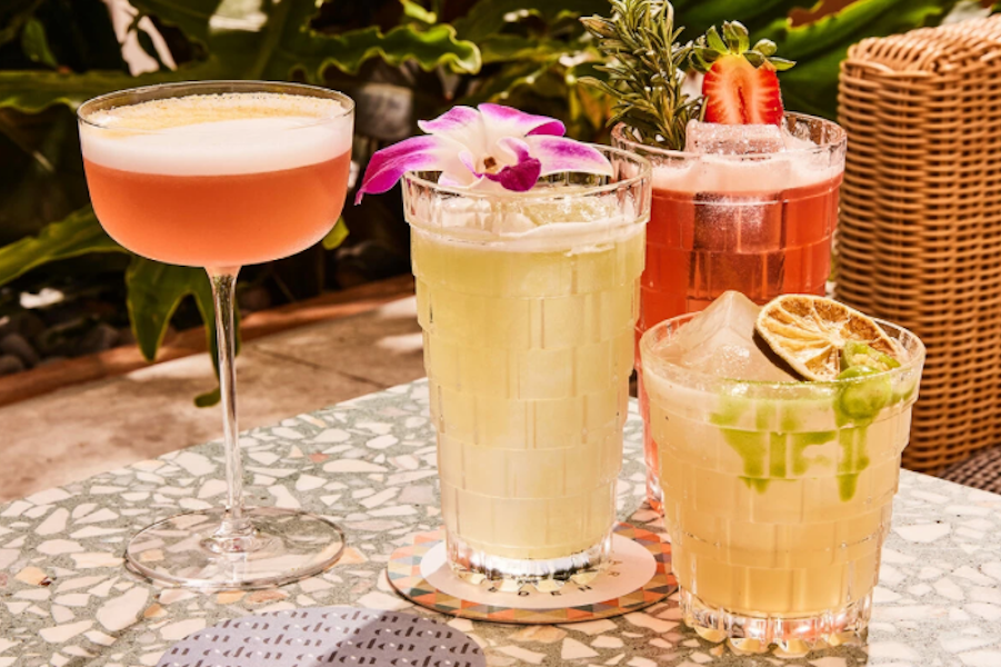 Assorted cocktails, including a pink drink in a coupe glass, a pale yellow drink with a purple flower, a red drink with a strawberry garnish, and a lime drink with dried lime slices, arranged on a patterned table outdoors.